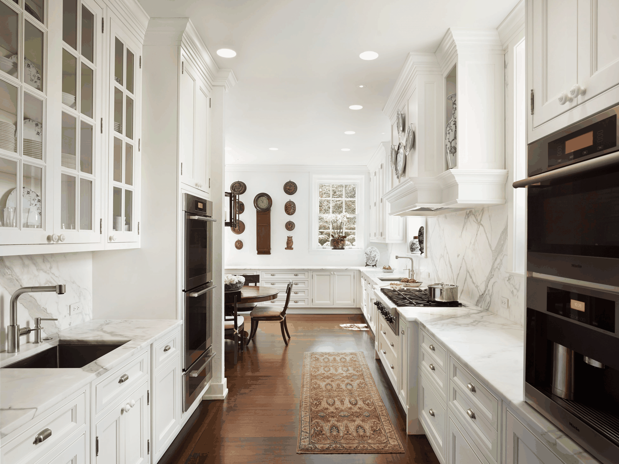 Bright white galley kitchen with marble backsplash, dark wood flooring and a patterned runner rug, with a dining area visible beyond, airy and clean mood