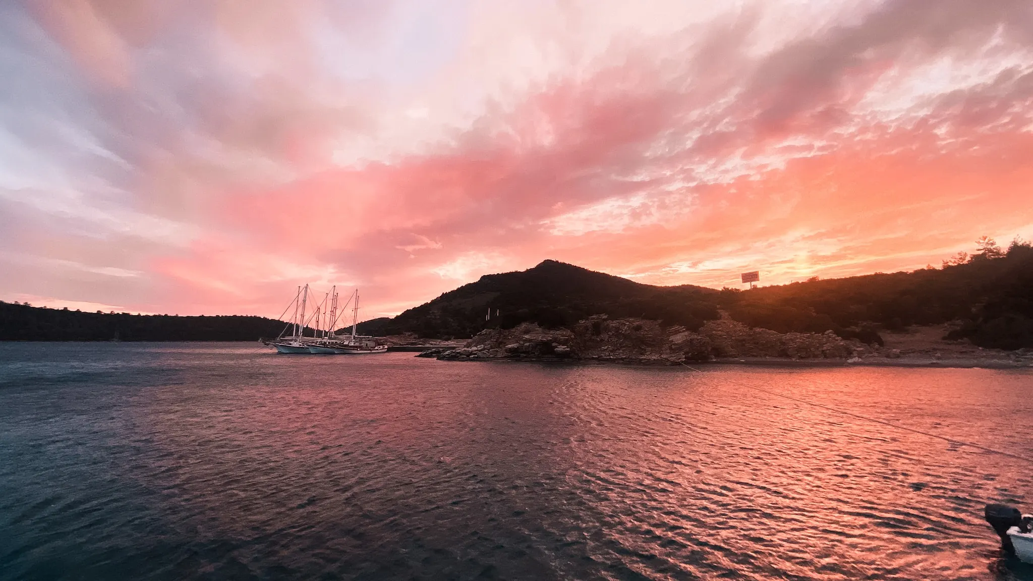 Sailboats resting on calm water near a rocky coastline under vivid pink and orange sunset clouds, dark hill silhouetted above